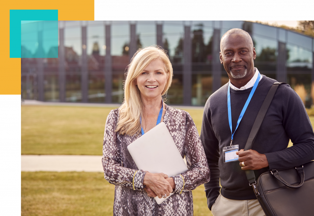 two college professors standing in front of a university building with nametags on lanyards holding a laptop