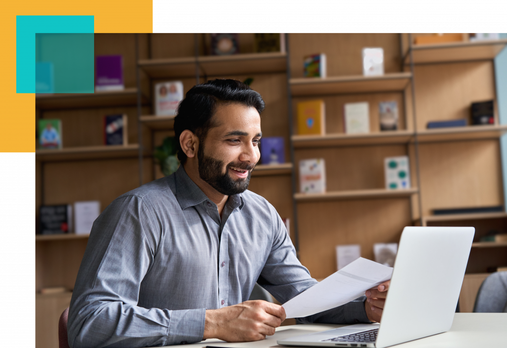 man smiling at a laptop as if on a conference or zoom call holding a sheet of paper