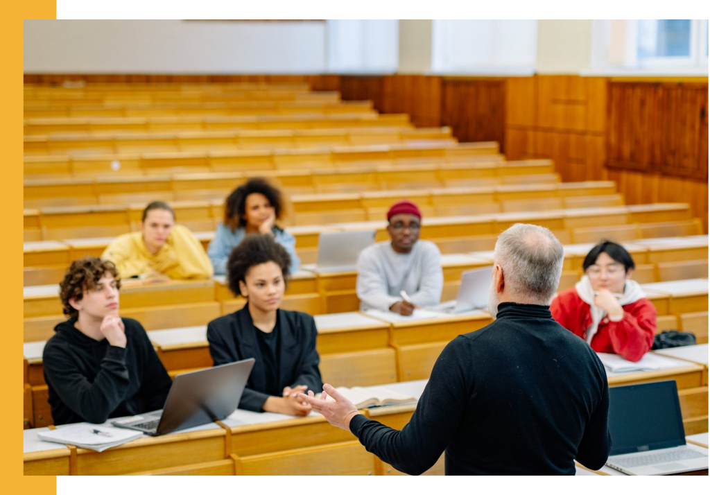 Professor teaching in a large college classroom, students listening with laptops in front of them